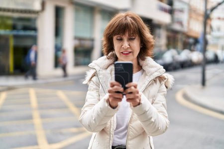 Téléchargez les photos : Femme d'âge moyen souriant confiant en utilisant smartphone à la rue - en image libre de droit