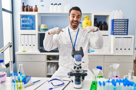 Photo for Young hispanic man with beard working at scientist laboratory looking confident with smile on face, pointing oneself with fingers proud and happy. - Royalty Free Image