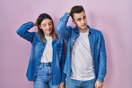 Foto de Young hispanic couple standing over pink background confuse and wondering about question. uncertain with doubt, thinking with hand on head. pensive concept. - Imagen libre de derechos