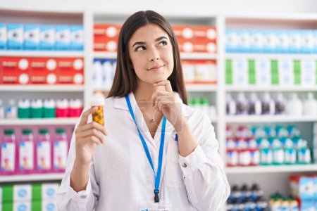Foto de Mujer hispana que trabaja en farmacia sosteniendo pastillas cara seria pensando en la pregunta con la mano en la barbilla, reflexivo acerca de la idea confusa - Imagen libre de derechos
