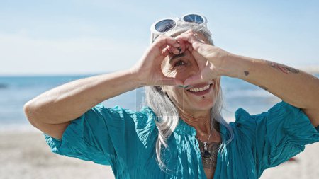 Foto de Turista mujer de pelo gris de mediana edad sonriendo confiado haciendo gesto de corazón con las manos en la playa - Imagen libre de derechos