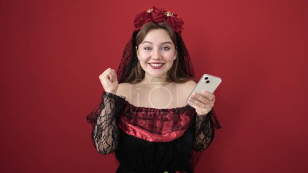 Téléchargez les photos : Jeune femme blonde portant un costume de katrina en utilisant un smartphone sur fond rouge isolé - en image libre de droit