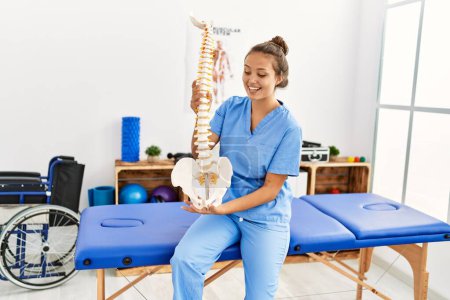 Téléchargez les photos : Jeune belle femme hispanique physiothérapeute souriant confiant tenant modèle anatomique de la colonne vertébrale à la clinique de réadaptation - en image libre de droit