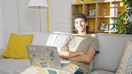 Foto de Joven hombre hispano usando laptop sentado en sofá con los brazos cruzados gesto en casa - Imagen libre de derechos