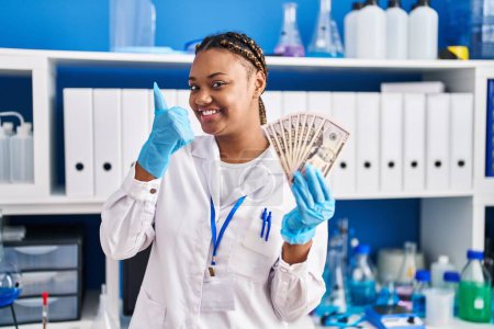 Photo for African american woman with braids working at scientist laboratory holding money smiling happy and positive, thumb up doing excellent and approval sign - Royalty Free Image