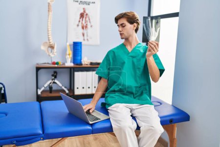 Foto de Joven hombre caucásico fisioterapeuta buscando rayos X utilizando ordenador portátil en la clínica de rehabilitación - Imagen libre de derechos