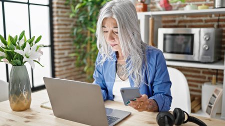 Photo for Middle age grey-haired woman using laptop and smartphone sitting on table at dinning room - Royalty Free Image