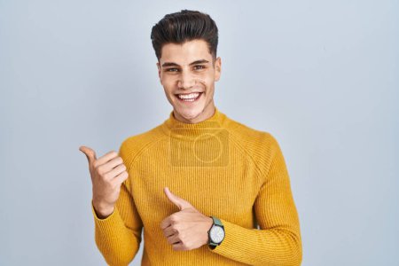 Téléchargez les photos : Young hispanic man standing over blue background pointing to the back behind with hand and thumbs up, smiling confident - en image libre de droit