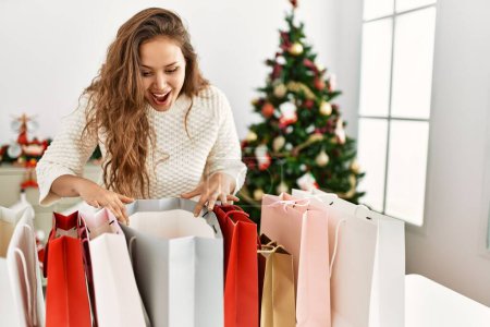 Téléchargez les photos : Jeune belle femme hispanique à la recherche de sacs à provisions debout près de l'arbre de Noël à la maison - en image libre de droit