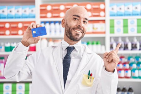 Téléchargez les photos : Moyen Age chauve homme travaillant à la pharmacie pharmacie tenant carte de crédit souriant heureux pointant avec la main et le doigt vers le côté - en image libre de droit