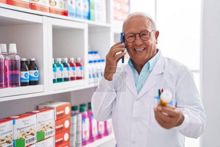 Téléchargez les photos : Homme âgé aux cheveux gris pharmacien parlant sur smartphone tenant des pilules à la pharmacie - en image libre de droit