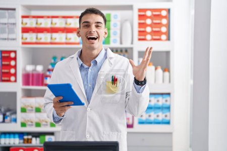 Téléchargez les photos : Bel homme hispanique travaillant à la pharmacie drugstore avec tablette célébrant la victoire avec sourire heureux et expression gagnante avec les mains levées - en image libre de droit