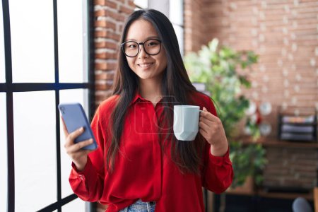 Foto de Young chinese woman business worker using smartphone drinking coffee at office - Imagen libre de derechos