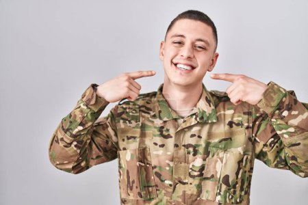 Téléchargez les photos : Jeune homme portant uniforme de camouflage de l'armée souriant gai montrant et pointant du doigt les dents et la bouche. concept de santé dentaire. - en image libre de droit