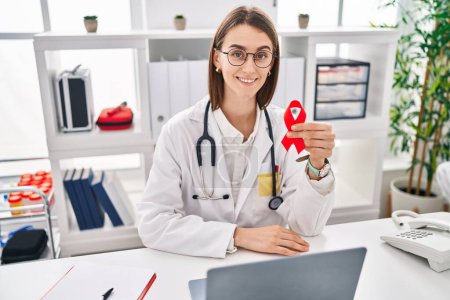 Photo for Young caucasian doctor woman holding support red ribbon looking positive and happy standing and smiling with a confident smile showing teeth - Royalty Free Image
