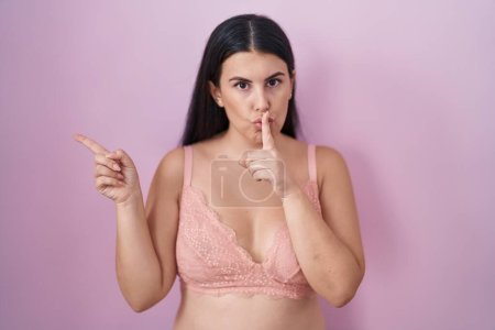 Photo for Young hispanic woman wearing pink bra asking to be quiet with finger on lips pointing with hand to the side. silence and secret concept. - Royalty Free Image