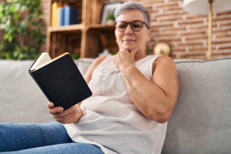 Photo for Middle age woman reading bible sitting on sofa at home - Royalty Free Image