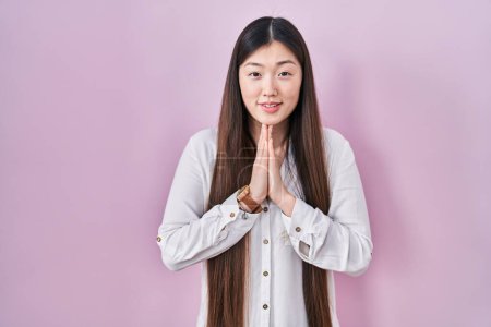 Téléchargez les photos : Chinois jeune femme debout sur fond rose priant avec les mains ensemble demandant pardon souriant confiant. - en image libre de droit