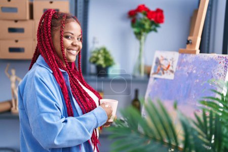 Téléchargez les photos : Femme artiste afro-américaine souriante boire du café au studio d'art - en image libre de droit