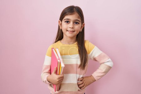 Photo for Little hispanic girl holding books looking positive and happy standing and smiling with a confident smile showing teeth - Royalty Free Image