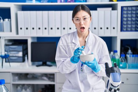 Foto de Mujer joven china trabajando en laboratorio científico mezclando en la cara de choque, mirando escéptico y sarcástico, sorprendido con la boca abierta - Imagen libre de derechos