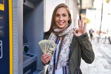 Foto de Joven mujer rubia sosteniendo billetes de dólares de cajero automático haciendo signo ok con los dedos, sonriendo gesto amistoso excelente símbolo - Imagen libre de derechos