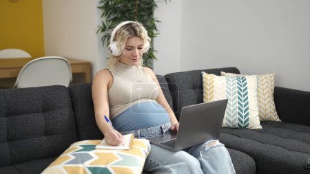 Photo for Young pregnant woman using laptop and headphones writing on notebook studying at home - Royalty Free Image