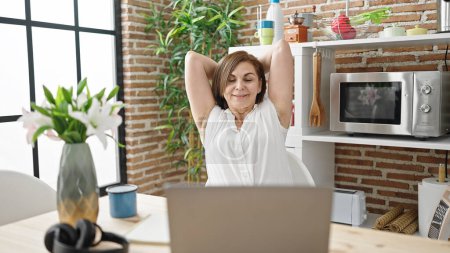 Photo for Middle age hispanic woman using laptop stretching arms at dinning room - Royalty Free Image