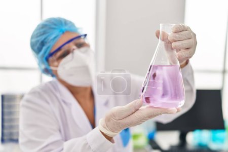 Téléchargez les photos : Femme d'âge moyen portant un uniforme scientifique et un masque médical tenant un tube à essai au laboratoire - en image libre de droit