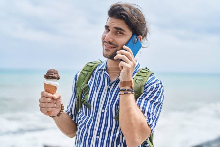 Téléchargez les photos : Jeune homme hispanique touriste parler sur smartphone manger de la crème glacée à la mer - en image libre de droit