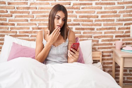 Photo for Young hispanic woman using smartphone sitting on the bed at home covering mouth with hand, shocked and afraid for mistake. surprised expression - Royalty Free Image