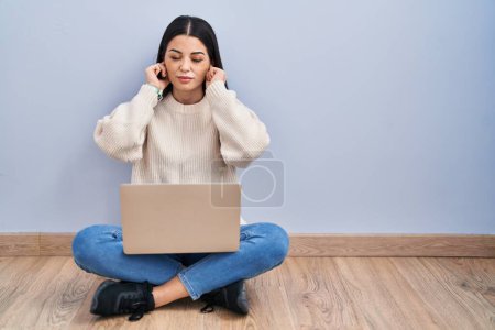 Téléchargez les photos : Jeune femme utilisant un ordinateur portable assis sur le sol à la maison couvrant les oreilles avec des doigts avec une expression agacée pour le bruit de la musique forte. concept sourd. - en image libre de droit