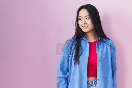 Photo for Young asian woman standing over pink background looking away to side with smile on face, natural expression. laughing confident. - Royalty Free Image
