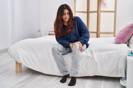 Foto de Mujer hispana joven que sufre de dolor de rodilla sentada en la cama en el dormitorio - Imagen libre de derechos