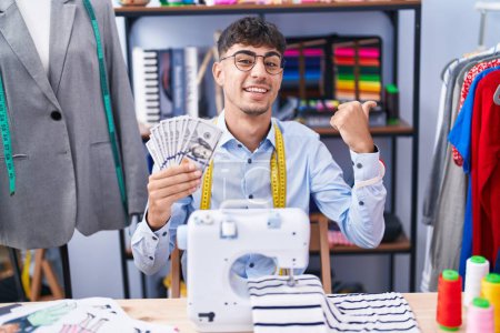 Photo for Young hispanic man dressmaker designer holding dollars pointing thumb up to the side smiling happy with open mouth - Royalty Free Image
