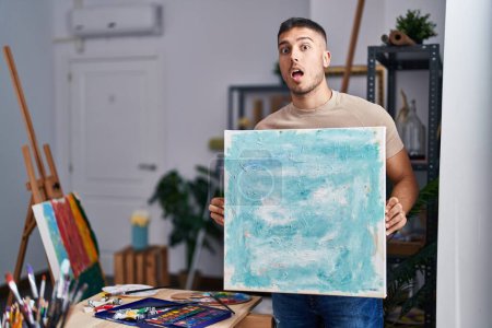 Photo for Young hispanic painter man holding canvas afraid and shocked with surprise and amazed expression, fear and excited face. - Royalty Free Image