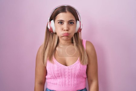 Foto de Mujer rubia joven escuchando música usando auriculares hinchando mejillas con cara divertida. boca hinchada de aire, expresión loca. - Imagen libre de derechos