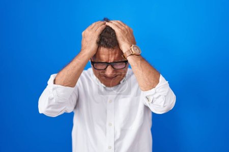 Photo for Middle age hispanic man standing over blue background suffering from headache desperate and stressed because pain and migraine. hands on head. - Royalty Free Image