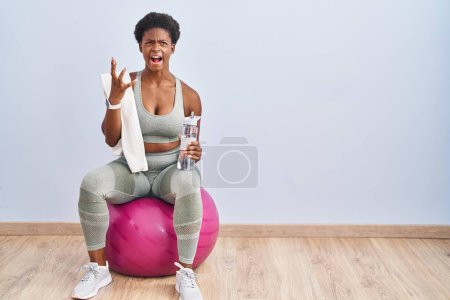 Photo for African american woman wearing sportswear sitting on pilates ball crazy and mad shouting and yelling with aggressive expression and arms raised. frustration concept. - Royalty Free Image