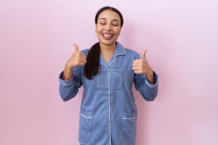 Foto de Mujer árabe joven vistiendo pijama azul signo de éxito haciendo gesto positivo con la mano, pulgares hacia arriba sonriendo y feliz. expresión alegre y gesto ganador. - Imagen libre de derechos