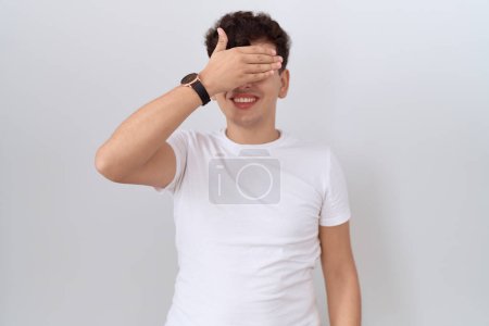 Téléchargez les photos : Jeune homme non binaire portant un t-shirt blanc décontracté souriant et riant avec la main sur le visage couvrant les yeux pour la surprise. concept aveugle. - en image libre de droit