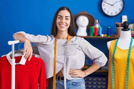 Téléchargez les photos : Jeune belle femme hispanique tailleur souriant confiant appuyé sur le porte-vêtements à l'usine de vêtements - en image libre de droit