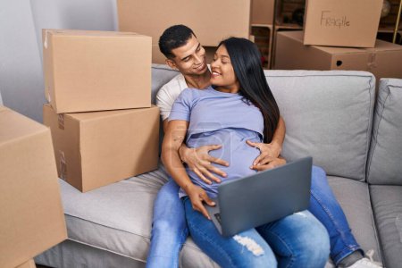 Photo for Young latin couple expecting baby touching belly using laptop at new home - Royalty Free Image