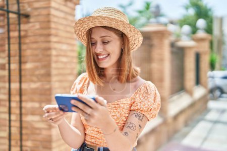 Foto de Joven mujer pelirroja turista con sombrero de verano viendo vídeo en el teléfono inteligente en la calle - Imagen libre de derechos