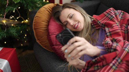 Photo for Young blonde woman using smartphone lying on sofa by christmas tree at home - Royalty Free Image
