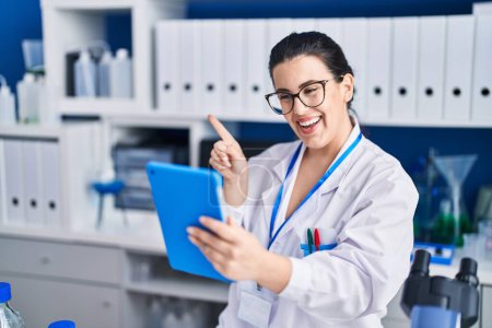Téléchargez les photos : Jeune femme brune travaillant au laboratoire scientifique en utilisant une tablette souriant heureux pointant avec la main et le doigt vers le côté - en image libre de droit