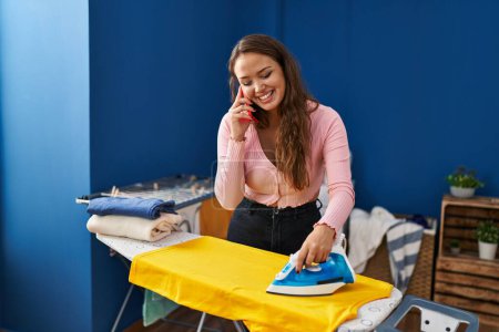 Foto de Joven hermosa mujer hispana hablando en smartphone planchando ropa en la lavandería - Imagen libre de derechos
