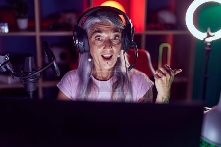 Photo for Middle age woman with tattoos playing video games wearing headphones pointing thumb up to the side smiling happy with open mouth - Royalty Free Image
