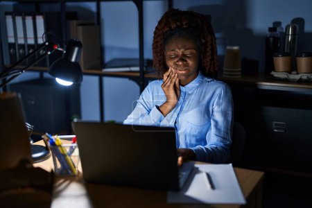 Téléchargez les photos : Femme africaine travaillant au bureau la nuit touchant la bouche avec la main avec une expression douloureuse à cause de maux de dents ou de maladies dentaires sur les dents. dentiste - en image libre de droit