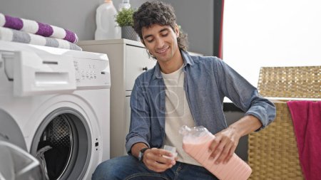 Téléchargez les photos : Jeune homme hispanique laver les vêtements verser détergent souriant à la buanderie - en image libre de droit